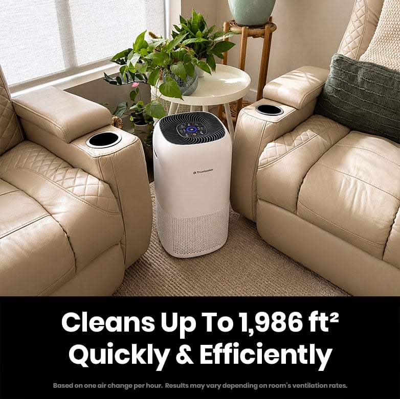 TrustedAir TruFlow smart HEPA air purifier placed between two beige leather recliners in a living room, with plants and natural light.
