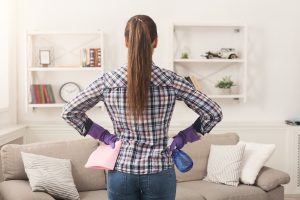 Woman cleaning living room with a spray bottle and cloth, wearing purple gloves, in a modern, tidy home.