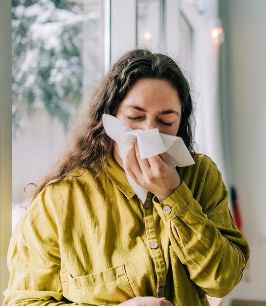 Woman with long curly hair using a tissue to blow her nose, indoors near a window.