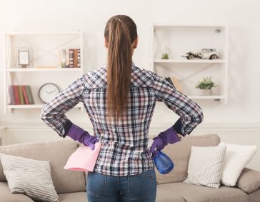 Woman cleaning living room with a spray bottle and cloth, wearing purple gloves, in a modern, tidy home.