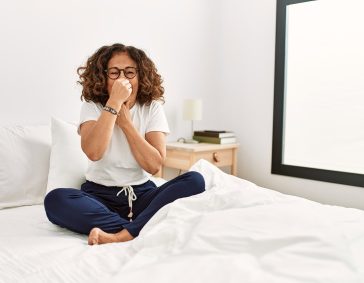 Woman sitting on bed covering her nose, demonstrating the use of an air purifier to reduce mold spores and improve indoor air quality.